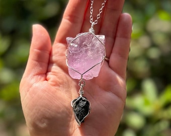 May include: A handcrafted necklace featuring a large, light purple amethyst crystal and a smaller black tourmaline crystal, both wrapped in silver-colored wire. The necklace is suspended from a delicate silver chain, showcasing the natural beauty of the stones.