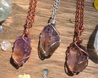 May include: Three necklaces with amethyst crystal pendants wrapped in copper wire. The necklaces are on a wooden surface with other crystals and flowers.