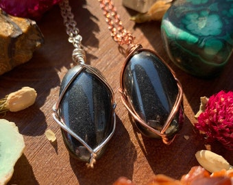 May include: Two silver and copper necklaces with black hematite stone pendants. The stones are wrapped in wire and have a smooth, polished finish. The necklaces are displayed on a wooden surface with other crystals and stones.