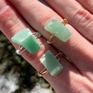 May include: Two rings featuring green gemstone pieces wrapped in gold and silver wire. The rings are worn on a person's finger.
