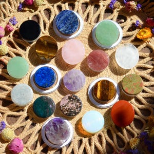 Puede incluir: Una colección de soportes circulares para teléfono con diversas piedras naturales pulidas en colores que incluyen azul, verde, rosa, marrón, morado, negro y blanco, dispuestos sobre una bandeja circular trenzada decorada con pequeñas flores secas.