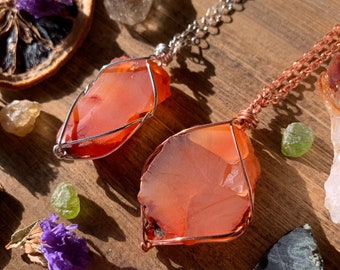 May include: Two carnelian crystal pendants wrapped in copper wire, hanging from silver and copper chains. The carnelian crystals are a vibrant orange-red color and have a natural, rough texture. The pendants are displayed on a wooden surface with other crystals and natural elements.