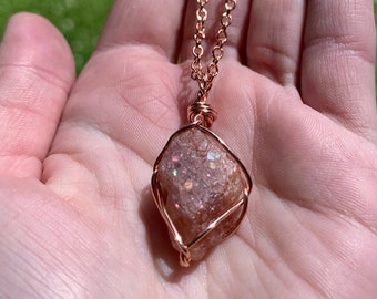May include: A copper wire wrapped pendant necklace featuring a raw, pink sunstone with iridescent sparkle.