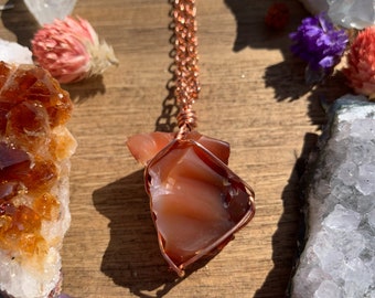 May include: A copper wire wrapped carnelian crystal pendant necklace. The carnelian crystal is a deep orange-red color and has a rough, natural shape. The copper chain is a simple, delicate design.