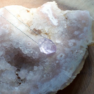May include: A silver chain necklace with a light purple amethyst crystal pendant. The pendant is resting on a white agate geode.