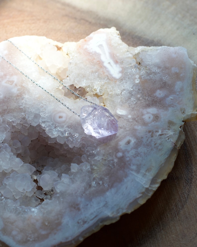 May include: A silver chain necklace with a light purple amethyst crystal pendant. The pendant is resting on a white agate geode.