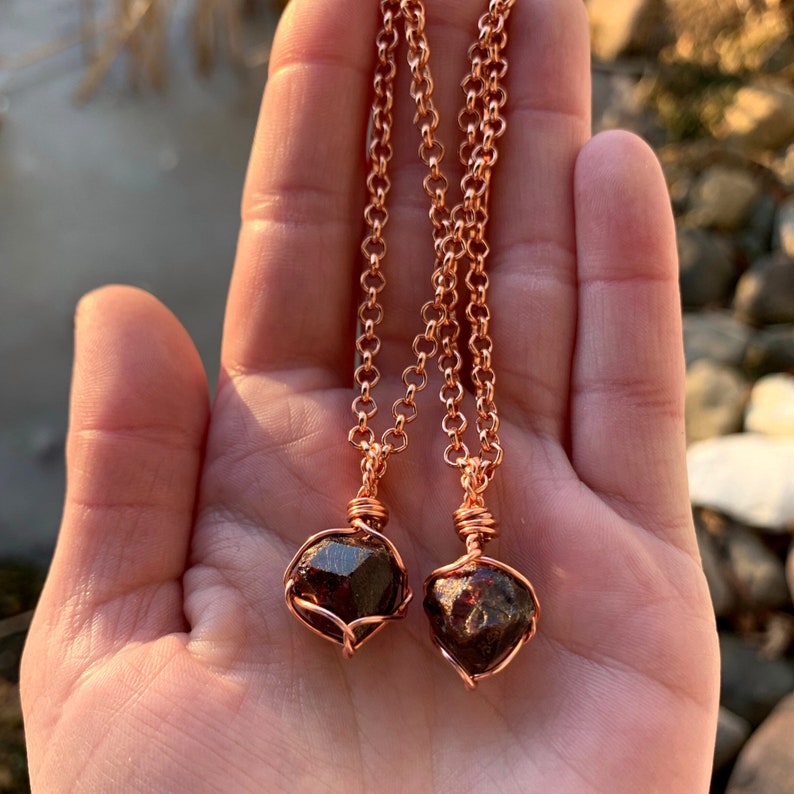 May include: Two copper necklaces with dark red garnet stones wrapped in copper wire. The necklaces are on a hand with a light brown background.