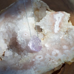 May include: A silver chain necklace with a light purple amethyst crystal pendant. The pendant is a rough, natural shape and is resting on a white and pink agate geode.