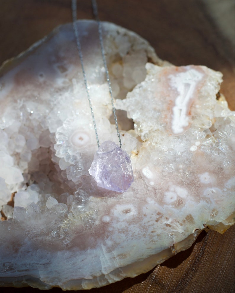 May include: A silver chain necklace with a light purple amethyst crystal pendant. The pendant is a rough, natural shape and is resting on a white and pink agate geode.