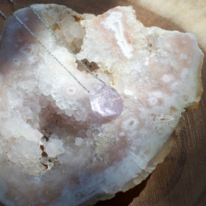 May include: A silver chain necklace with a light purple amethyst crystal pendant. The pendant is a geometric shape and is resting on a white and pink agate geode.