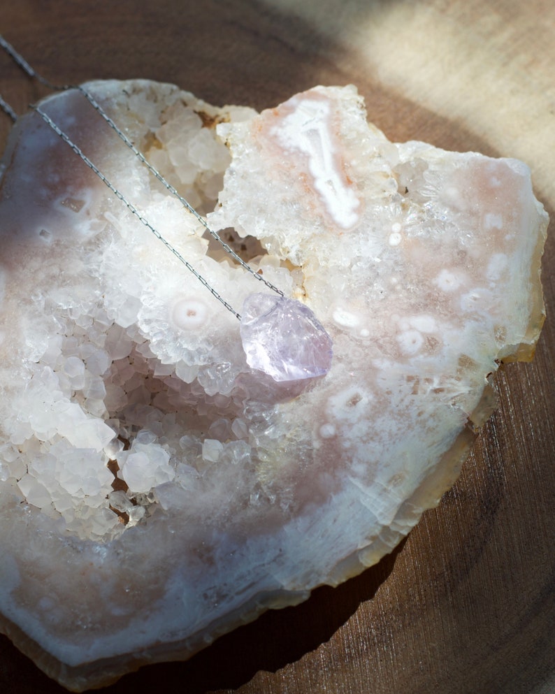 May include: A silver chain necklace with a light purple amethyst crystal pendant. The pendant is a geometric shape and is resting on a white and pink agate geode.