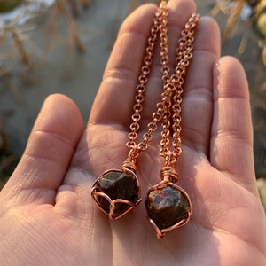 May include: Two copper necklaces with raw garnet stones wrapped in copper wire. The necklaces are on a hand with a blurred background.