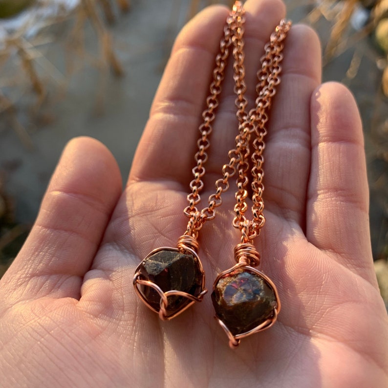 May include: Two copper necklaces with raw garnet stones wrapped in copper wire. The necklaces are on a hand with a blurred background.