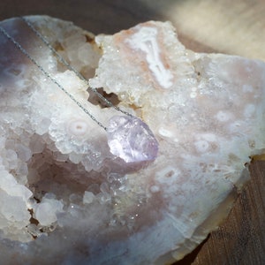 May include: A delicate silver chain necklace with a light purple amethyst crystal pendant. The pendant is a natural, rough-cut stone with a unique shape. The necklace is displayed on a white, textured surface.