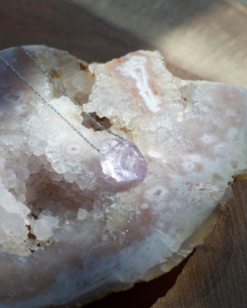 May include: A delicate silver chain necklace with a light purple amethyst crystal pendant. The pendant is a natural, rough-cut stone with a unique shape. The necklace is displayed on a white, textured surface.