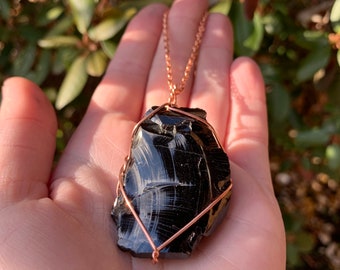 Könnte beinhalten: Eine Halskette mit Anhänger aus schwarzem Obsidian mit einer kupferfarbenen Kette. Der Obsidianstein ist unregelmäßig geformt und mit Kupferdraht umwickelt. Der Hintergrund ist verschwommen mit grünem Laub.