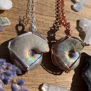 May include: Two fossil ammonite pendants. One is wrapped in silver wire on a silver chain, the other in copper wire on a copper chain. The fossils have iridescent colors and are set against a wooden background.