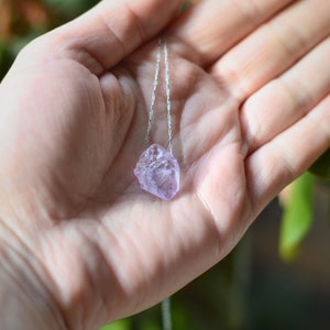 May include: A delicate silver chain necklace with a light purple amethyst crystal pendant. The crystal is raw and has a natural, irregular shape.