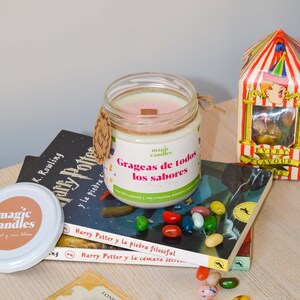 Puede incluir: Un tarro de vela blanco con tapa rosa y una etiqueta que dice "Grageas de todos los sabores". La vela está sentada sobre una pila de libros de Harry Potter en español. También hay gominolas de colores esparcidas alrededor de la vela.