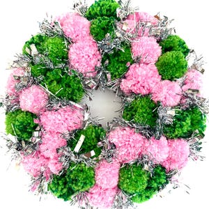 May include: A round wreath composed of pink and green pom-poms, interspersed with silver tinsel. The wreath is a festive decoration, suitable for various holidays or seasonal displays. The pom-poms are densely packed, creating a full and textured appearance.