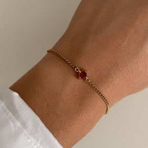 Peut inclure: Un bracelet en chaîne dorée avec un petit pendentif en forme de carré de pierre rouge.