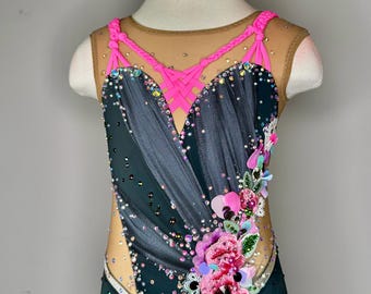 May include: A black and grey figure skating costume with pink and multi-colored floral embellishments. The costume has a sweetheart neckline and a mesh bodice with a criss-cross design. The costume also has a black skirt with a V-shaped panel in the front.