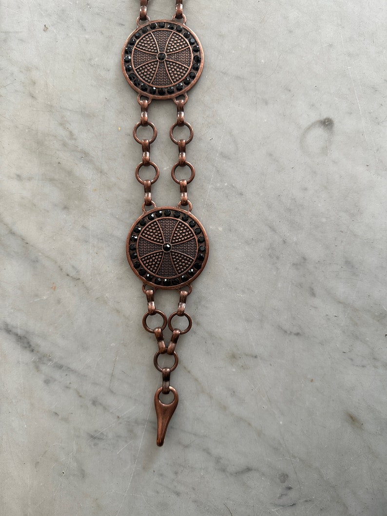 May include: A copper chain belt with two round black and copper medallions. Each medallion has a cross design with black rhinestones.