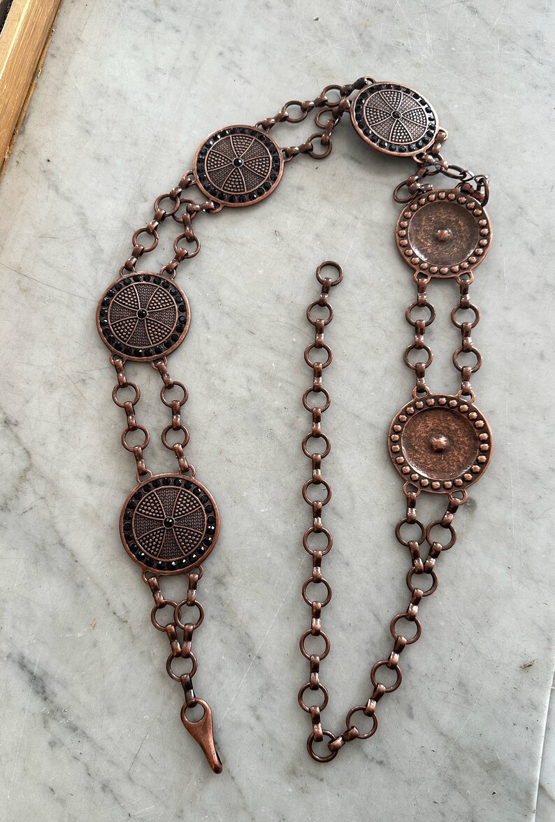May include: A copper chain belt with five round, decorative medallions. The medallions have a black rhinestone design and a textured surface.