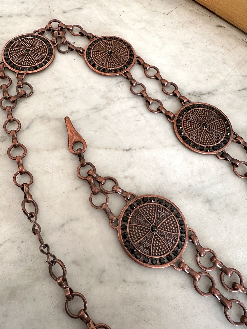 May include: A copper chain belt with four round, decorative, black rhinestone-encrusted medallions.