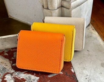 May include: Three small leather wallets in orange, yellow, and white. The wallets are stacked on top of each other, with the orange wallet on the bottom.