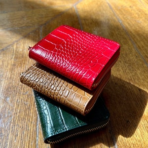 Può includere: Tre piccoli portafogli con cerniera, impilati uno sull'altro, di colore rosso, marrone e verde. I portafogli hanno un motivo strutturato a forma di pelle di coccodrillo. Su una superficie di legno.