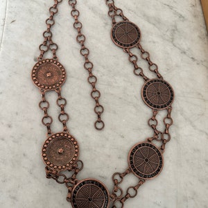 May include: A copper chain belt with five circular pendants. The pendants have a black center with a geometric design and a copper border.