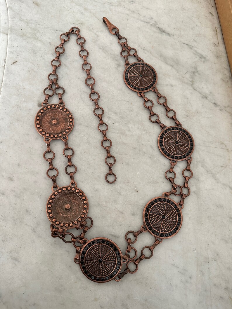 May include: A copper chain belt with five circular pendants. The pendants have a black center with a geometric design and a copper border.