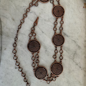 May include: A copper chain belt with five round, decorative, black-and-copper medallions. The medallions have a cross-like design.