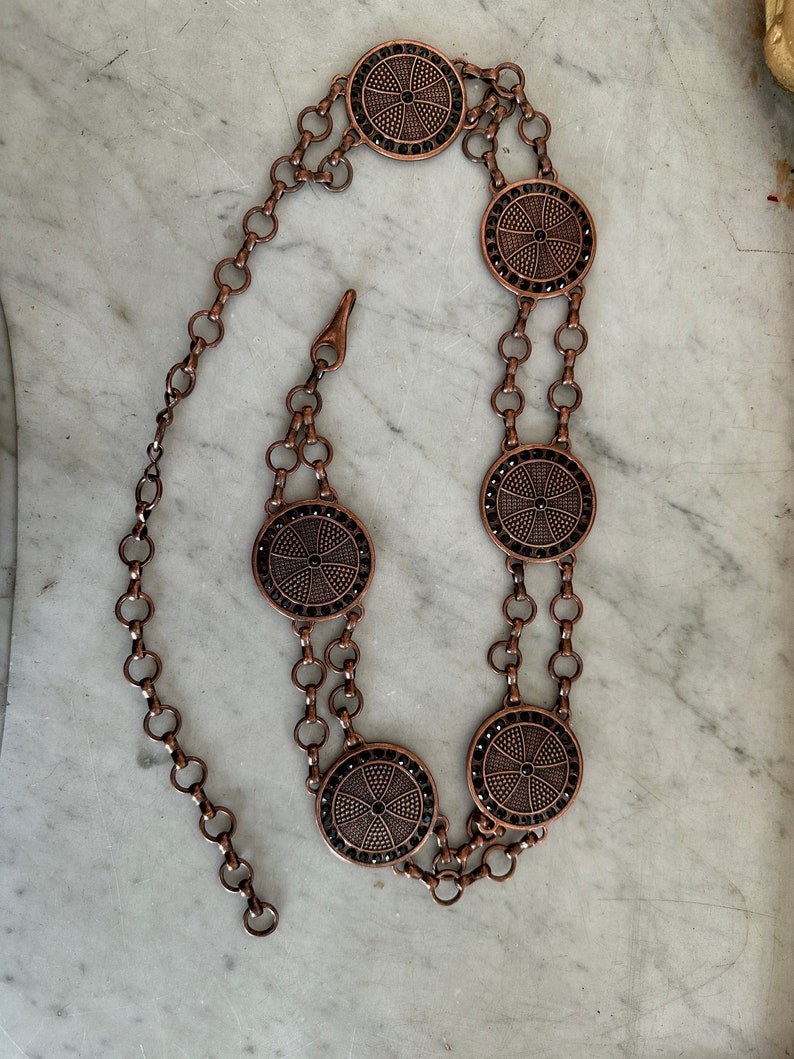 May include: A copper chain belt with five round, decorative, black-and-copper medallions. The medallions have a cross-like design.
