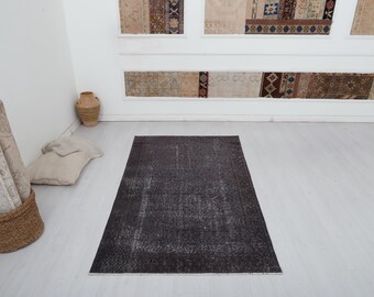 Puede incluir: Una alfombra rectangular gris oscuro con un aspecto envejecido y vintage. La alfombra está colocada sobre un suelo de madera de color claro. En el fondo se ven alfombras decorativas, junto con una cesta de mimbre y un jarrón de cerámica.