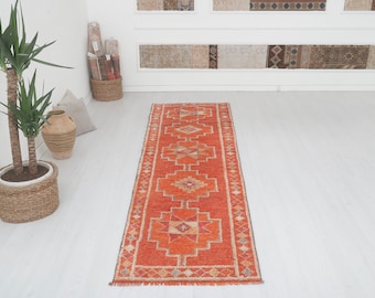 May include: A long, narrow, red and orange runner rug with a geometric pattern. The rug has a fringe on both ends.