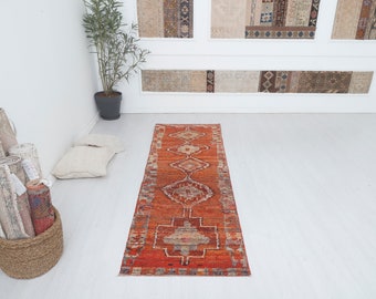 May include: A red, orange, and cream colored runner rug with a geometric pattern. The rug has a fringed edge and is laid out on a white floor.