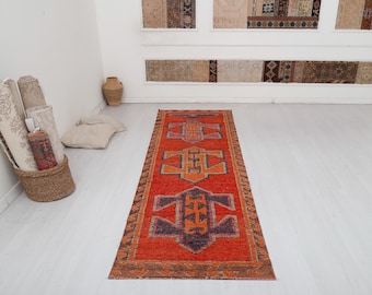 May include: A long, rectangular, red and orange rug with a geometric pattern. The rug has a dark purple and orange center design with a lighter orange border.