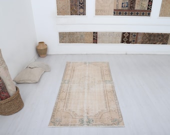 May include: A rectangular, beige rug with a detailed border and floral accents. The rug is placed on a light-colored floor, with a woven basket and rolled rugs visible in the background. The rug's design features a central field and ornate edges.