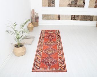 May include: A red, orange, and cream colored runner rug with a geometric pattern. The rug has a fringe border and is laid out on a white floor.