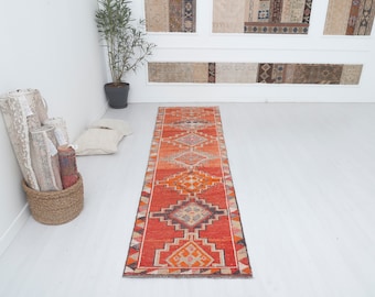 May include: A long, narrow red and orange runner rug with a geometric pattern. The rug has a traditional design with intricate details and a faded look.