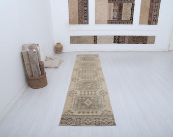 May include: A rectangular, beige and brown patterned runner rug. The rug features a repeating geometric design with a central medallion and border details. The rug is displayed on a light-colored floor, with rolled rugs and decorative items in the background.