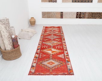 May include: A red, orange, and white patterned runner rug with geometric designs. The rug has a fringed edge and is laid out on a white floor.