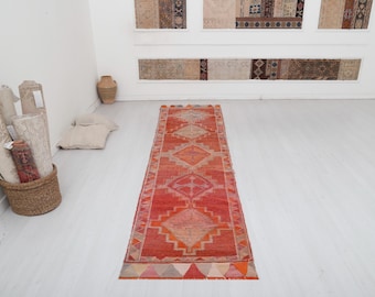 May include: A red, orange, and cream colored runner rug with a geometric pattern. The rug has a fringe border and is laid out on a white floor.