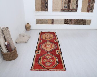 May include: A rectangular, red and orange patterned rug with geometric designs. The rug has a black border and is displayed on a white floor. Rolled rugs and a basket are in the corner.