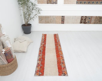 May include: A long, narrow, cream-colored rug with red and orange geometric patterns along the edges. The rug is laid out on a white floor.