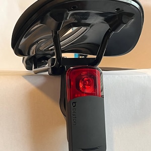 May include: A black bike seat with a Bryton brand rear bike light attached to it. The light has a red lens and a black body.