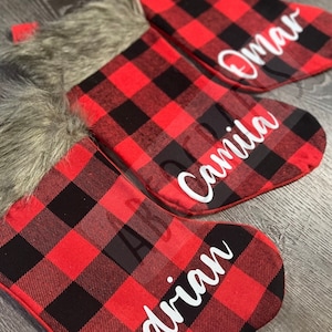 May include: Three red and black plaid Christmas stockings with white faux fur trim. The stockings are personalized with the names "Adrian", "Camila", and "Omar".