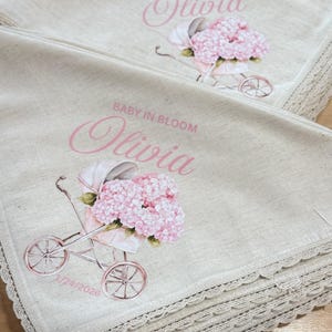 May include: Two beige baby blankets with a pink floral design and the text "Baby in Bloom" and "Olivia". Each blanket features a vintage baby carriage with pink flowers and a lace trim.